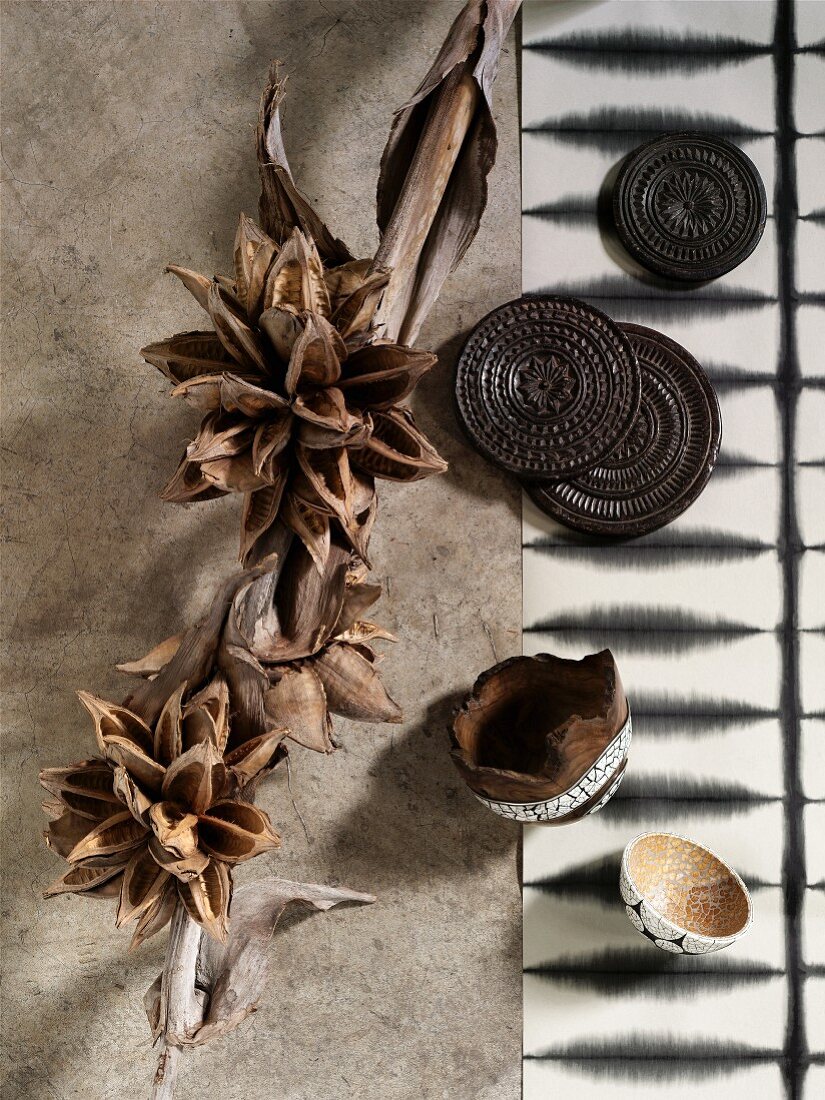 Getrocknete Samenschalen und Pflanzenteile neben kunstvoll bearbeiteten, afrikanischen Holzuntersetzern und schwarzweissem Batik-Läufer