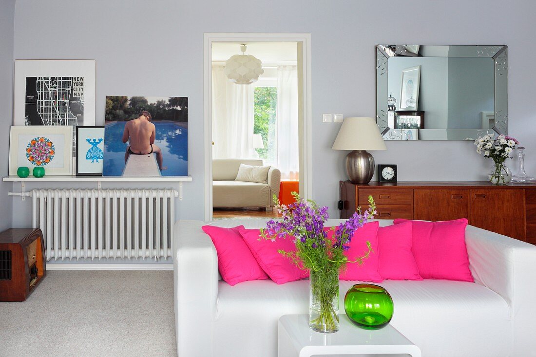 Weisses Polstersofa mit aneinandergereihten, pinkfarbenen Kissen, davor Beistelltisch mit Blumenvase in Wohnraum
