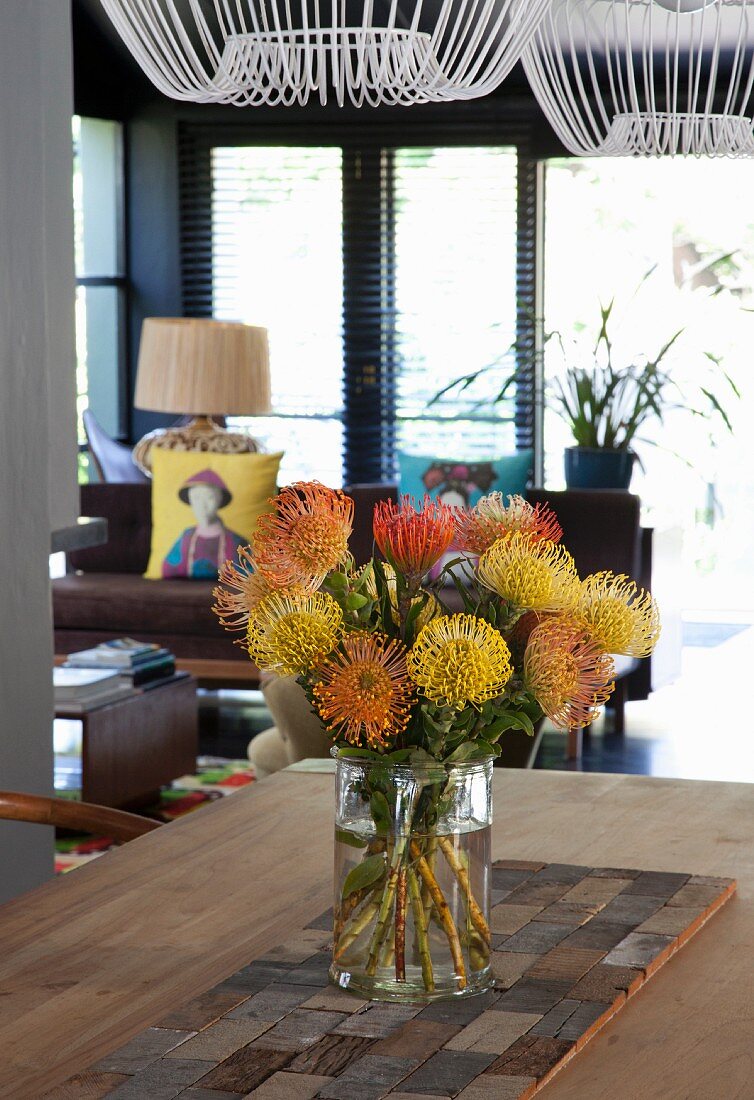 Tropical bouquet of pincushion proteas in glass vase on table; lounge area in background