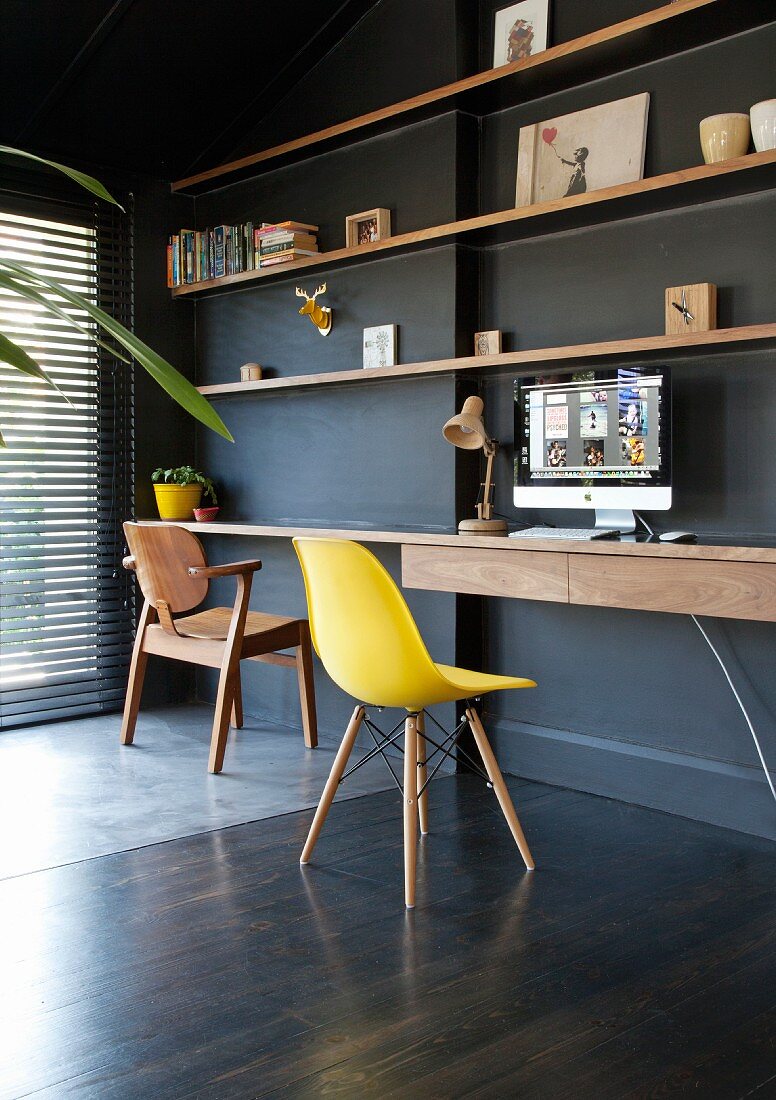 Gelber Klassiker Schalenstuhl vor minimalistischem Schreibtisch unter Regalböden aus Holz an schwarzer Wand