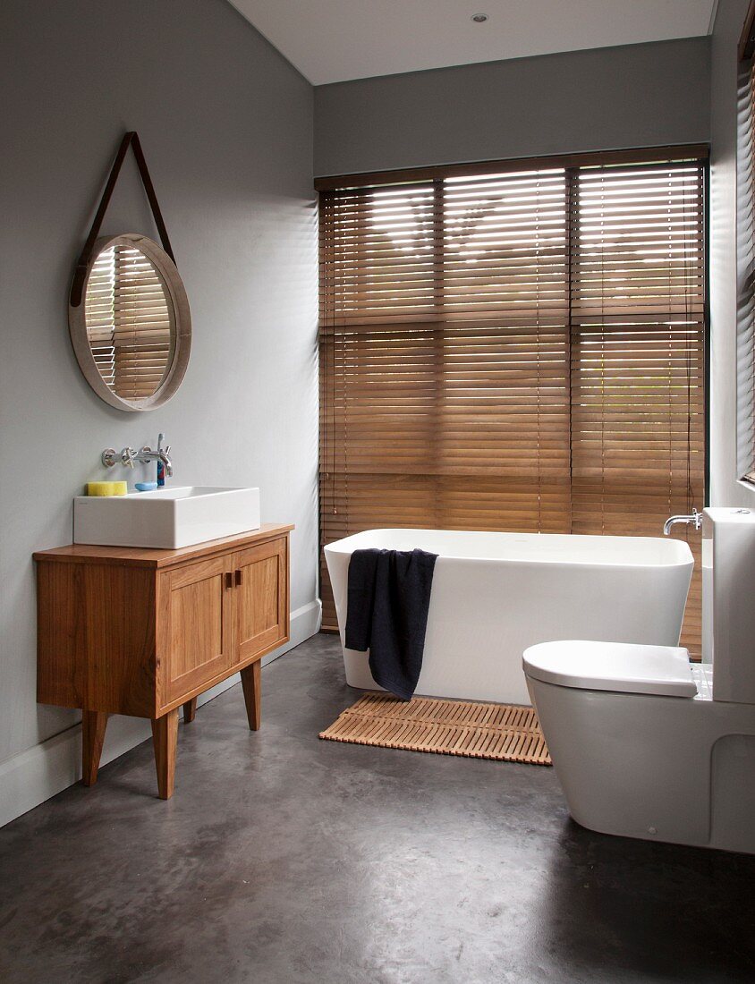Modernes Bad mit freistehender Badewanne vor Fenster und geschlossenr Jalousie, seitlich Waschtischmöbel aus Massivholz