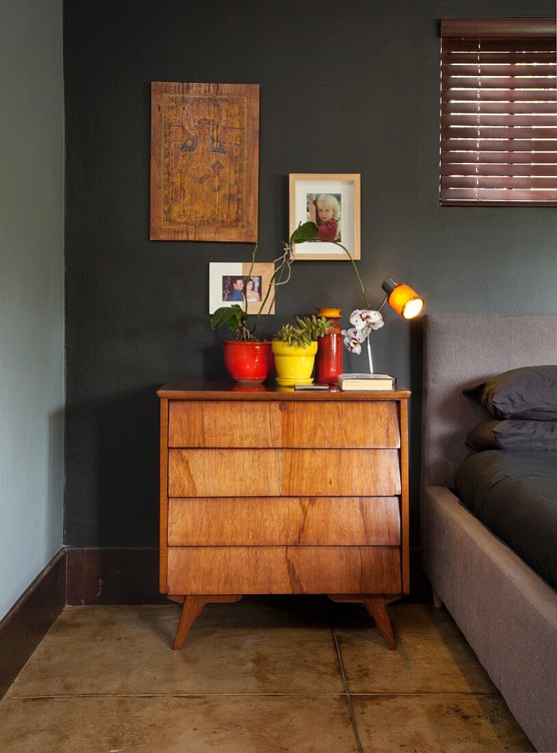 Colourful plant pots and retro table lamp on fifties-style, solid wood chest of drawers in bedroom with black-painted wall