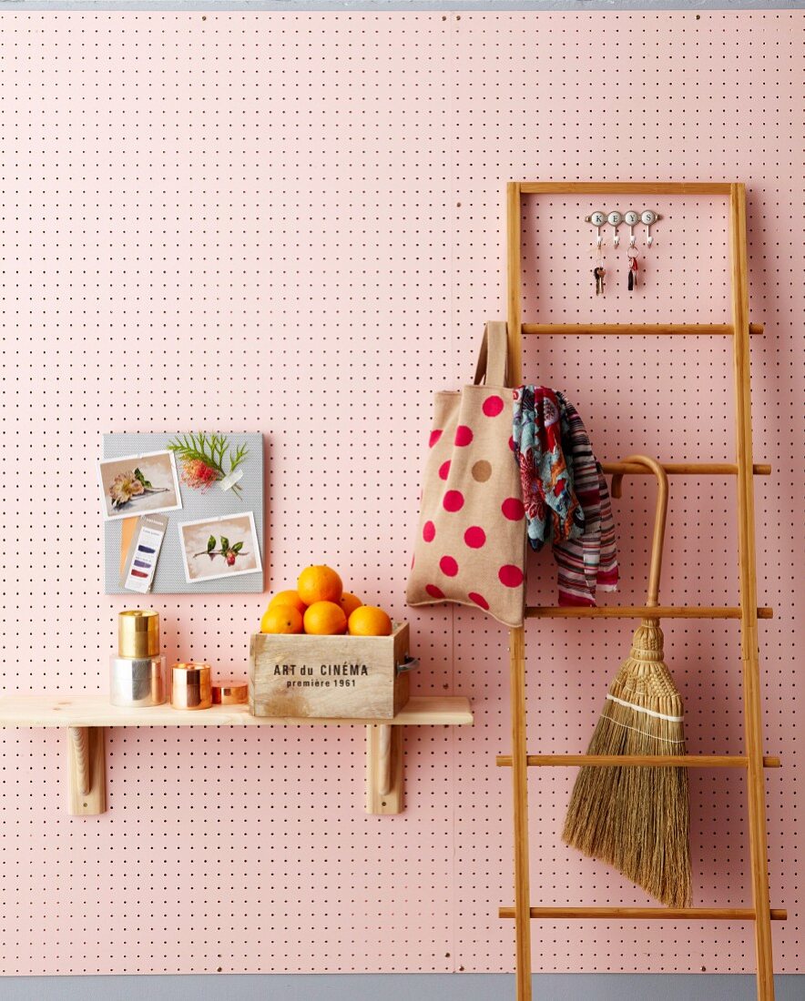Cloth shopping bag and hand brush hung on wooden ladder next to box of oranges on bracket shelf on pink perforated wall