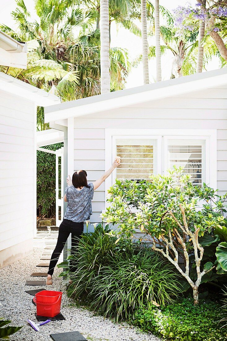 Woman cleaning windows - small, white wooden house with small front garden in tropical surroundings