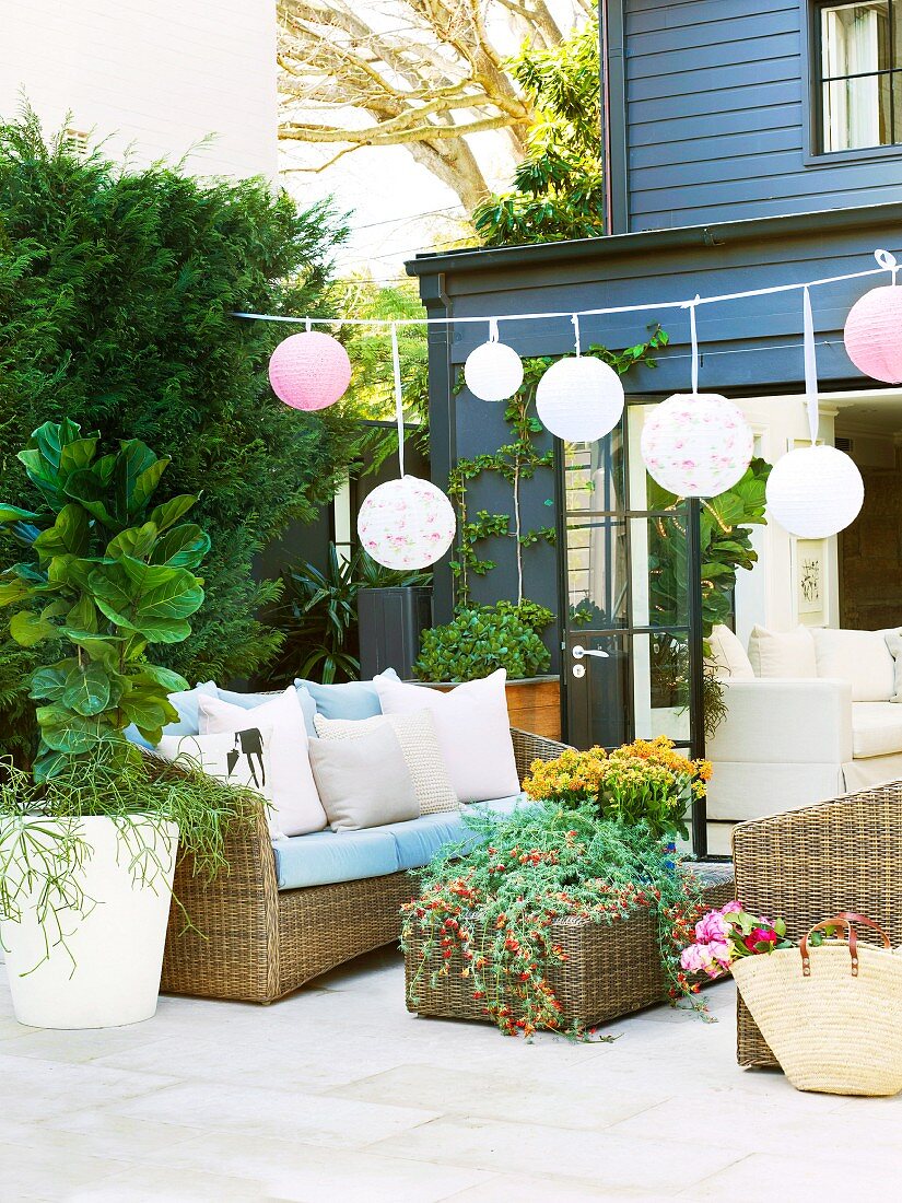 Garland of lanterns and rattan sofa set with flower arrangement on terrace of contemporary house with dark grey weatherboard facade