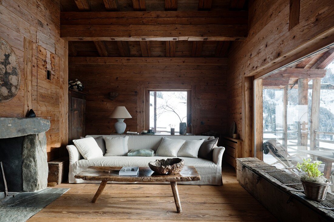 Pale, loose-covered sofa with scatter cushions behind rustic wooden coffee table in living room of cabin with panoramic window to one side