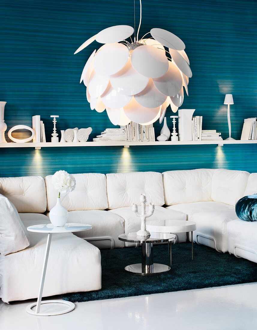 Seating area with white furnishings - designer pendant lamp with overlapping, fish-scale lampshade above sofa combination below wall-mounted shelf with spotlights on blue striped wall