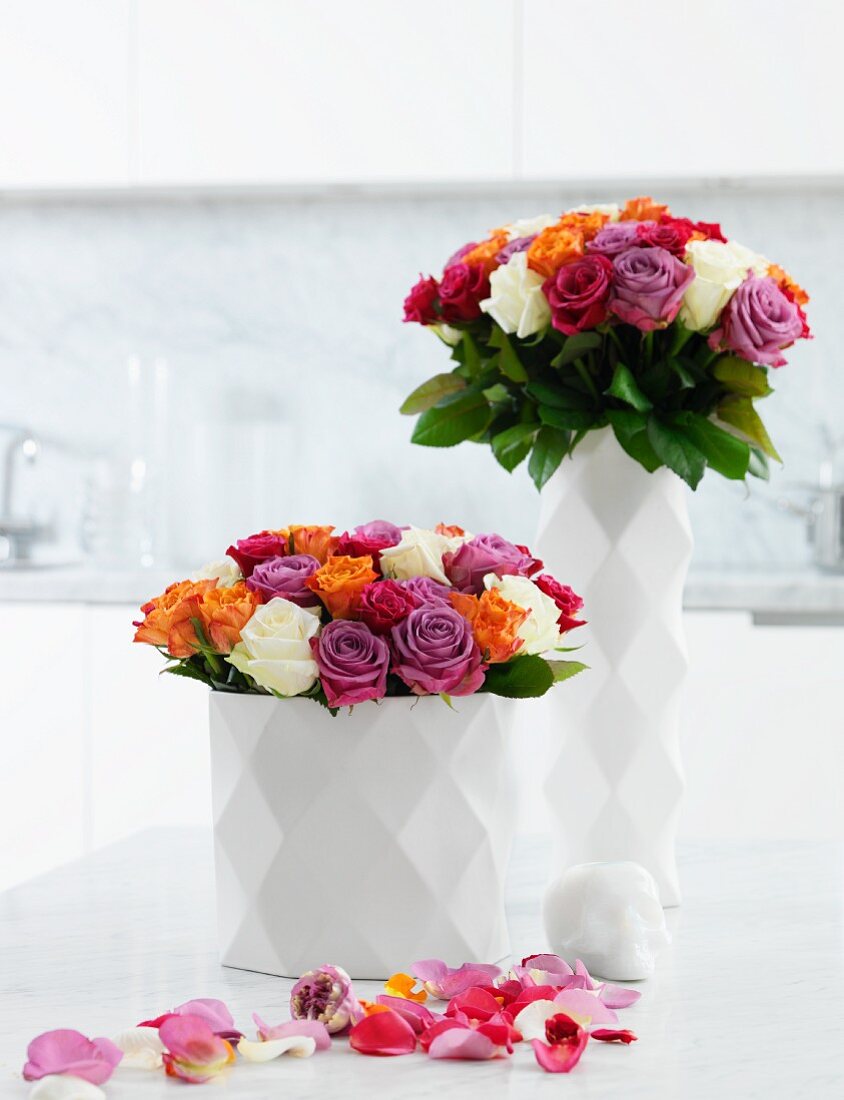 Multicoloured roses in white china vases with diamond-patterned structure surface and petals strewn on white surface