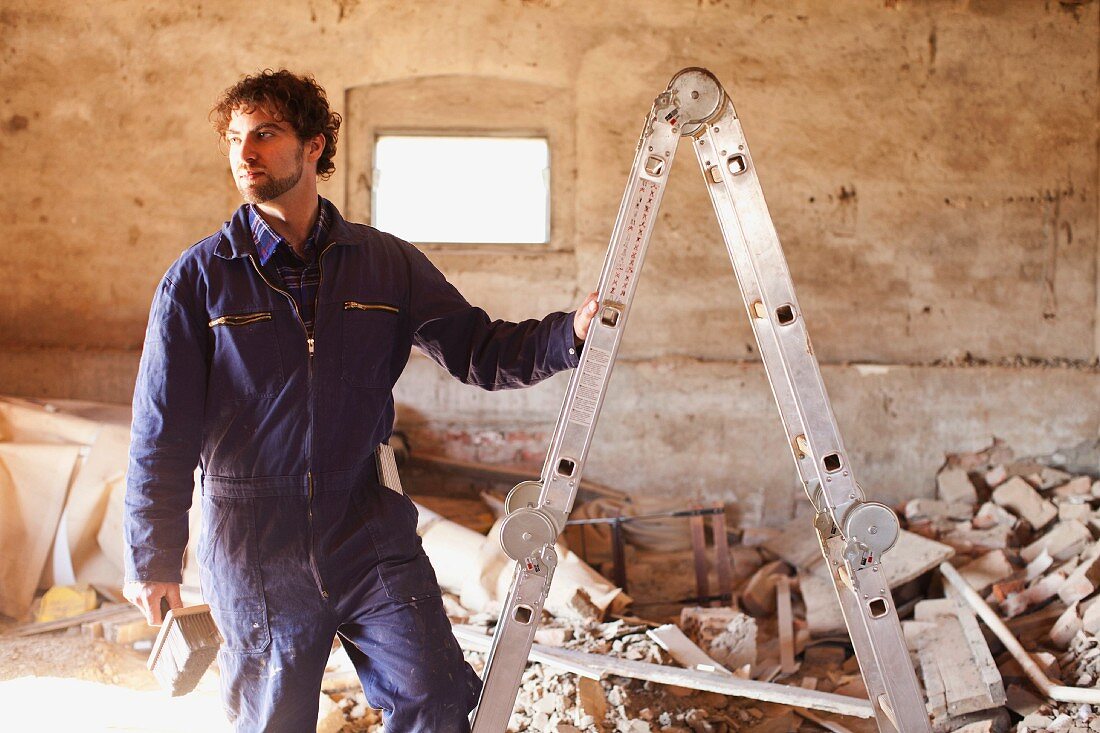 Mann im blauen Arbeitsanzug an Aluleiter in Raum mit Betonwand und Bauschutt