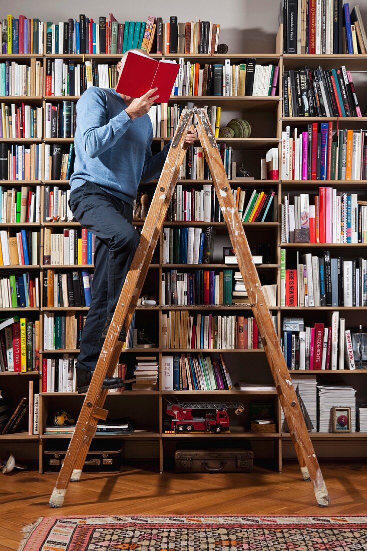 Mann liest Buch auf Malerleiter vor Bücherwand