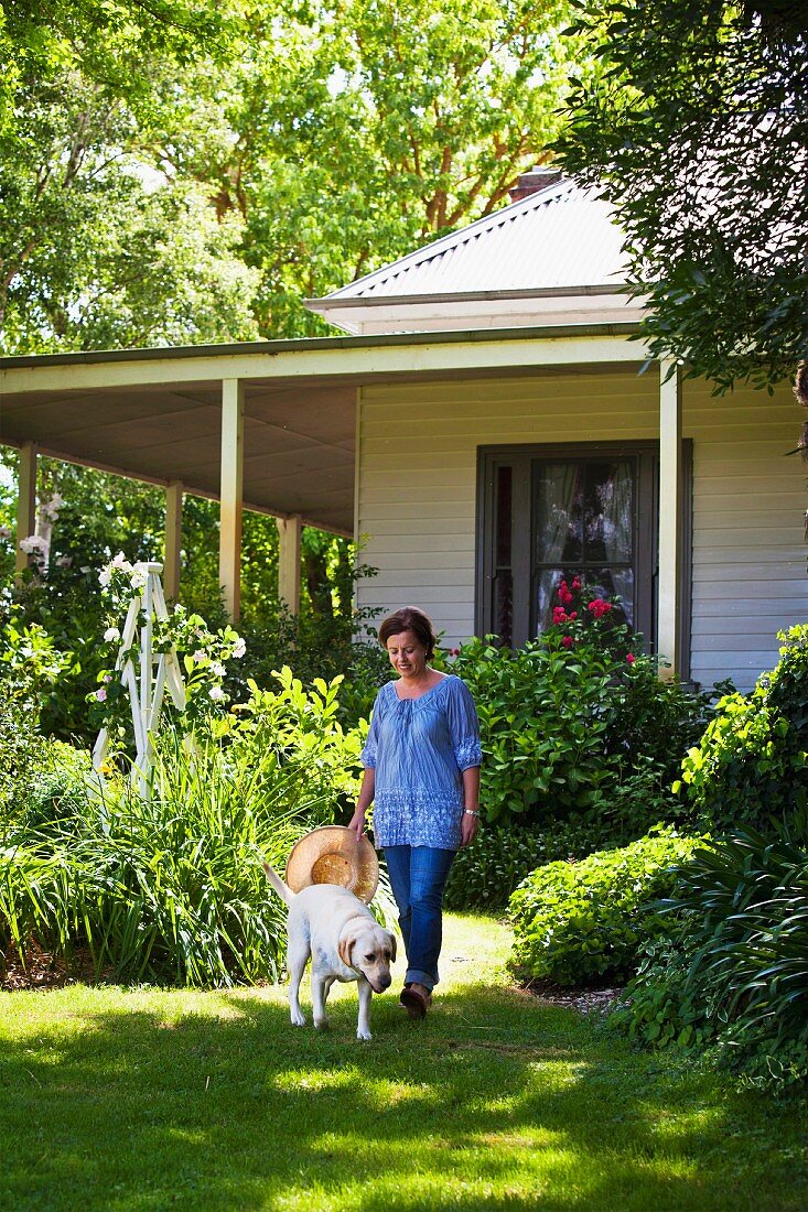 Frau mit Hund im sommerlichen Garten vor holzverkleidetem weißen Landhaus mit Laubengang