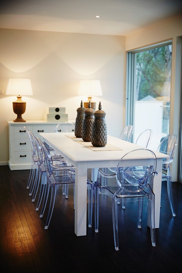 Speisezimmer mit weißem Holztisch und Plexiglasstühlen, Schubladenkommode und Lampen