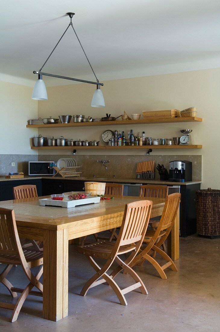 Solid wooden dining table in minimalist, country-house kitchen with open, floating shelves above kitchen worksurface