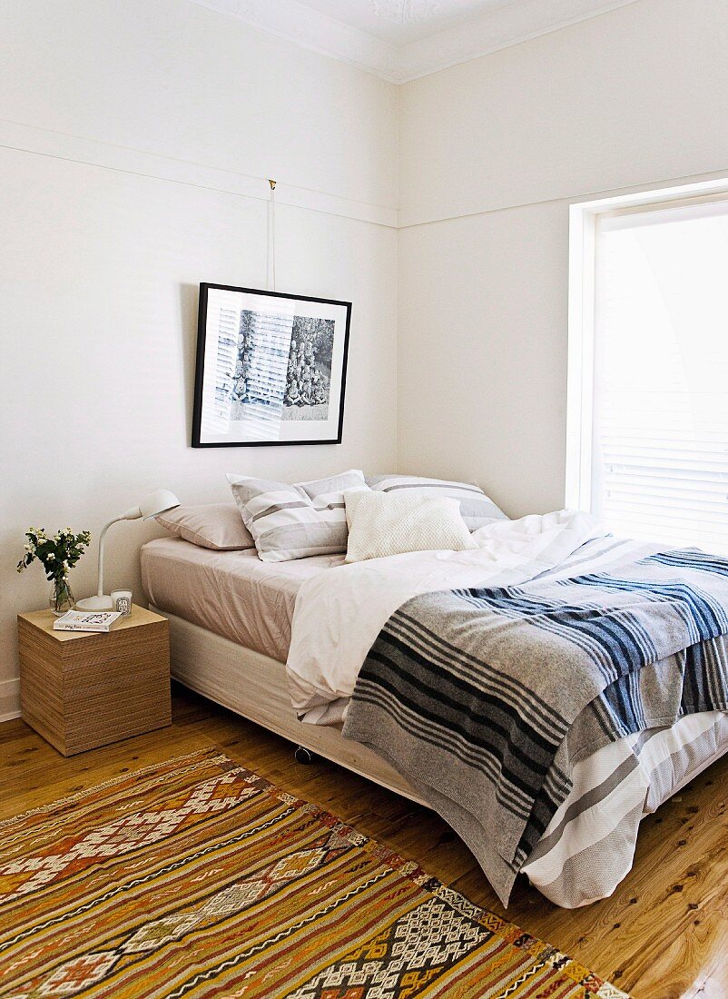 Folkloristischer Teppich neben Bett mit gestreifter Tagesdecke in schlichtem Schlafzimmer