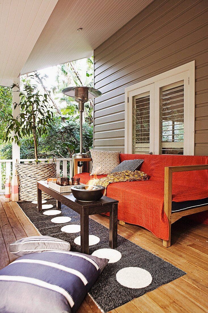 A wooden coffee table on a long rug with a circular pattern in front of a bench with an orange throw on the veranda of a wooden house
