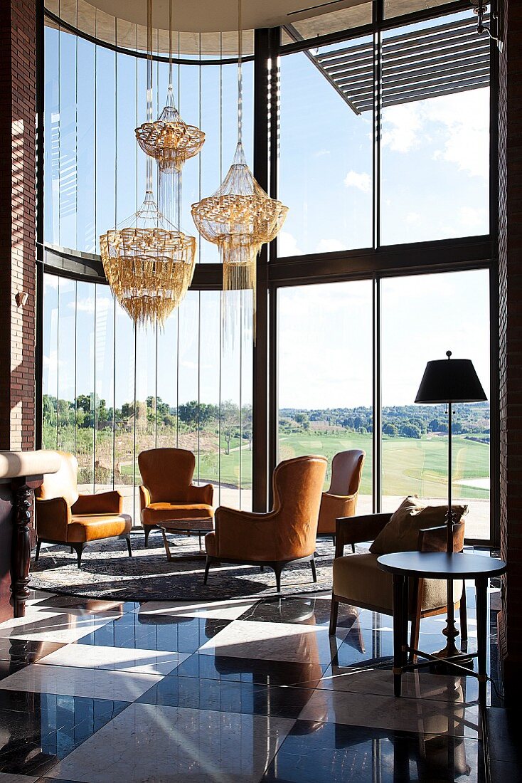 Zeitgenössisches Clubhaus mit gebogener Glasfassade und verschieden hoch gehängte Kronleuchter über Sitzgruppen im Salon mit Aussicht