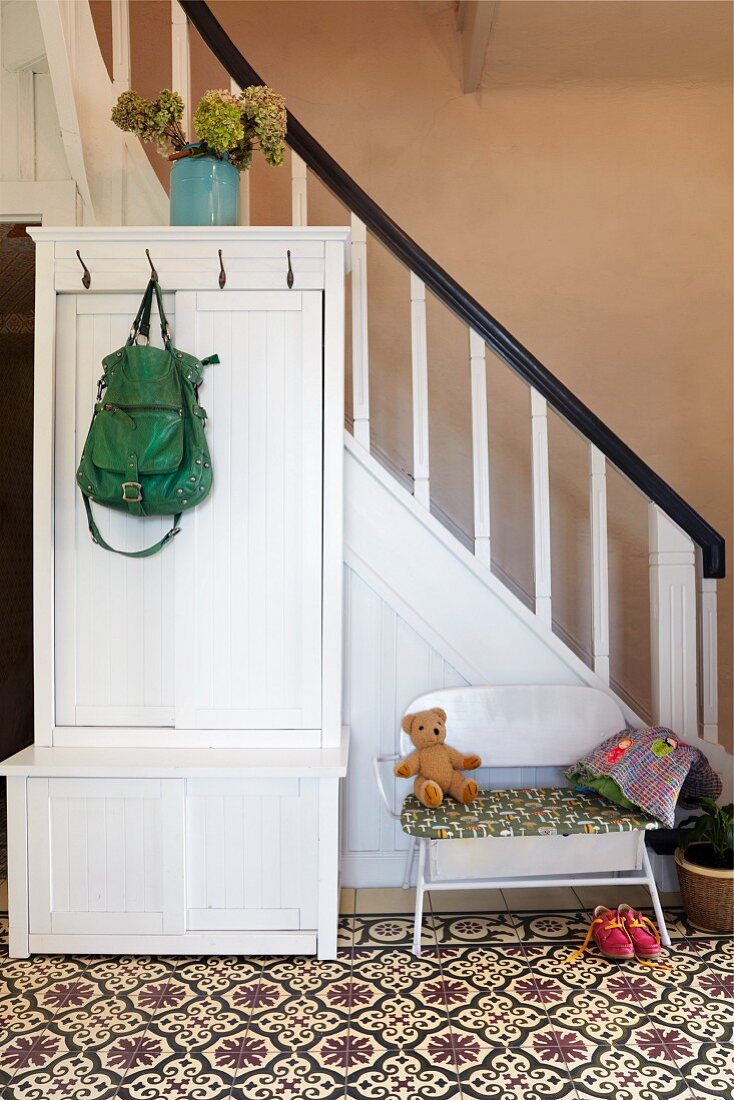 Garderobenschrank aus weiss lackiertem Holz vor Treppenaufgang in rustikalem Hausflur mit gemustertem Fliesenboden