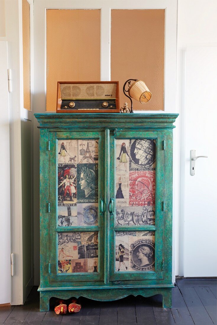Green cabinet with door panels covered in newspaper clippings in corner of room below transom window