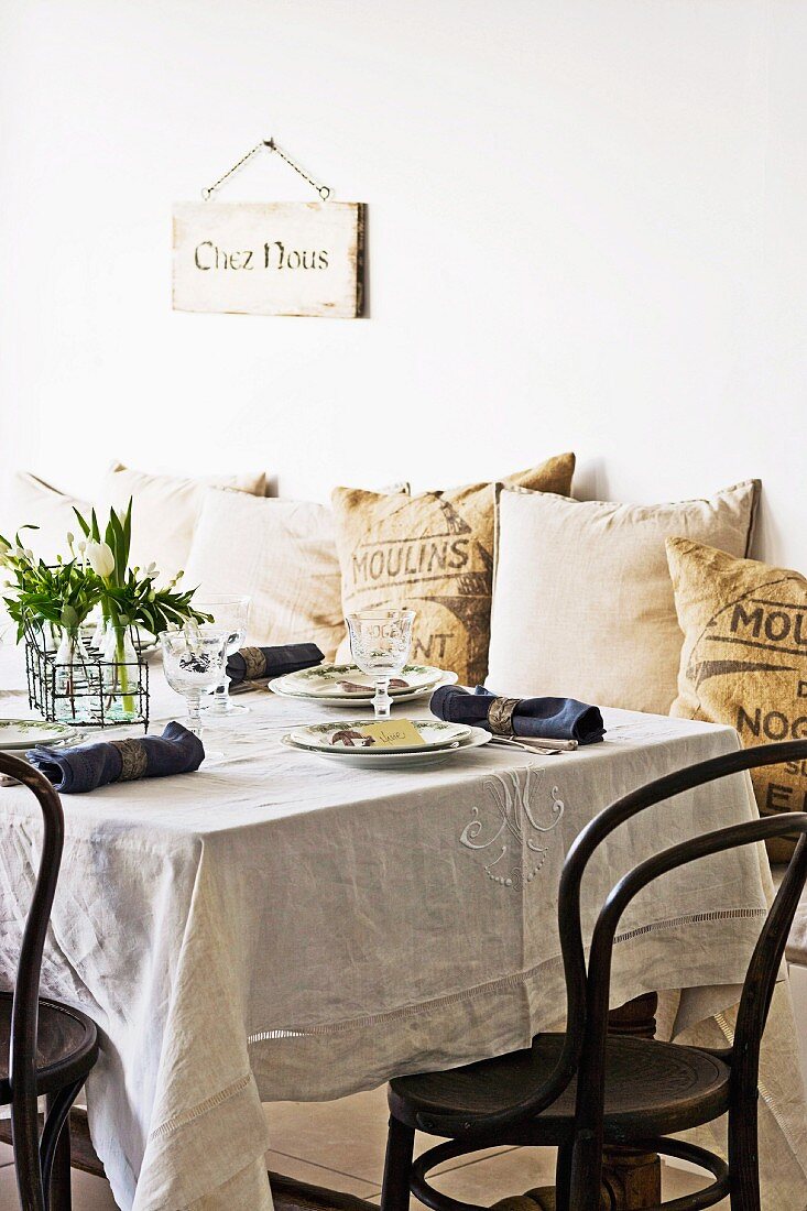 Esstischdeko im französischen Stil mit bestickter Tischdecke und Thonetstühlen; im Hintergrund Kissen aus Sackleinen mit Aufdruck und Wandschild