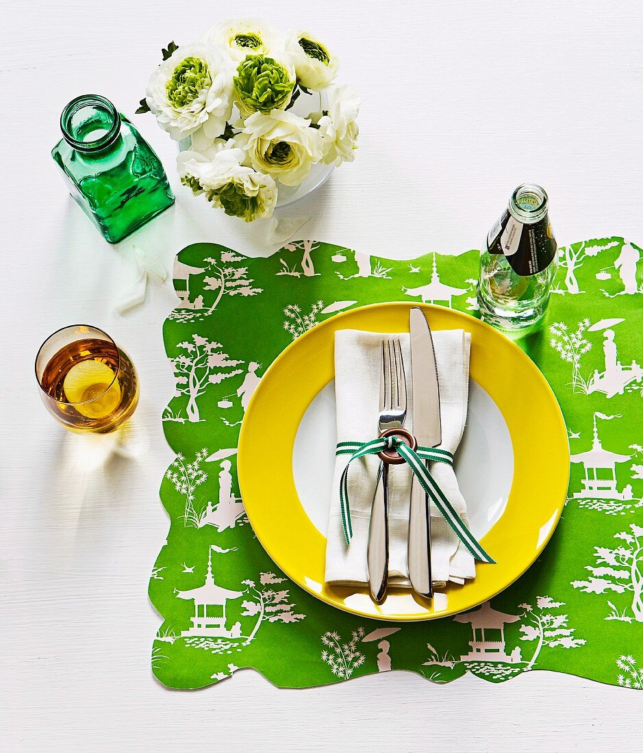 Besteck und Serviette mit Geschenkband verschnürt auf Teller mit gelbem Rand und Set aus bedrucktem Geschenkpapier mit Wellenrand