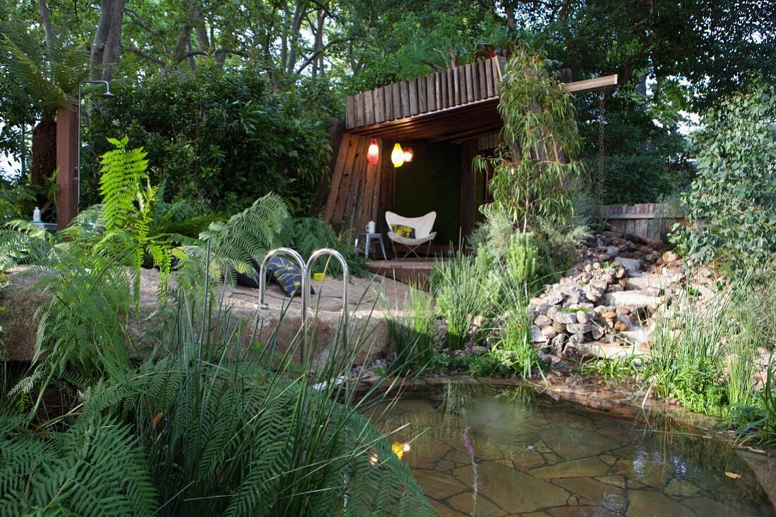 Pool mit Natursteinen und Bepflanzung, im Hintergrund ein Badehaus mit Butterfly Chair