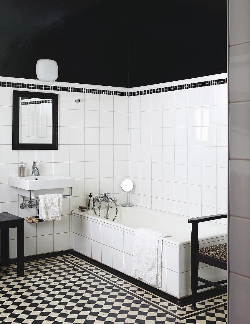 Badezimmer mit schwarzweissem Fliesenboden, Waschbecken und Badewanne vor weissen Wandfliesen, darüber schwarze Wand