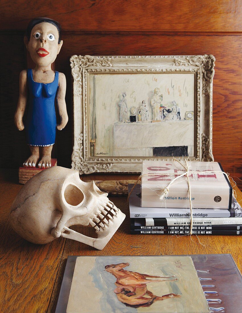 Still-life arrangement of fake skull, stacked books, painted wooden figurine and picture on table top