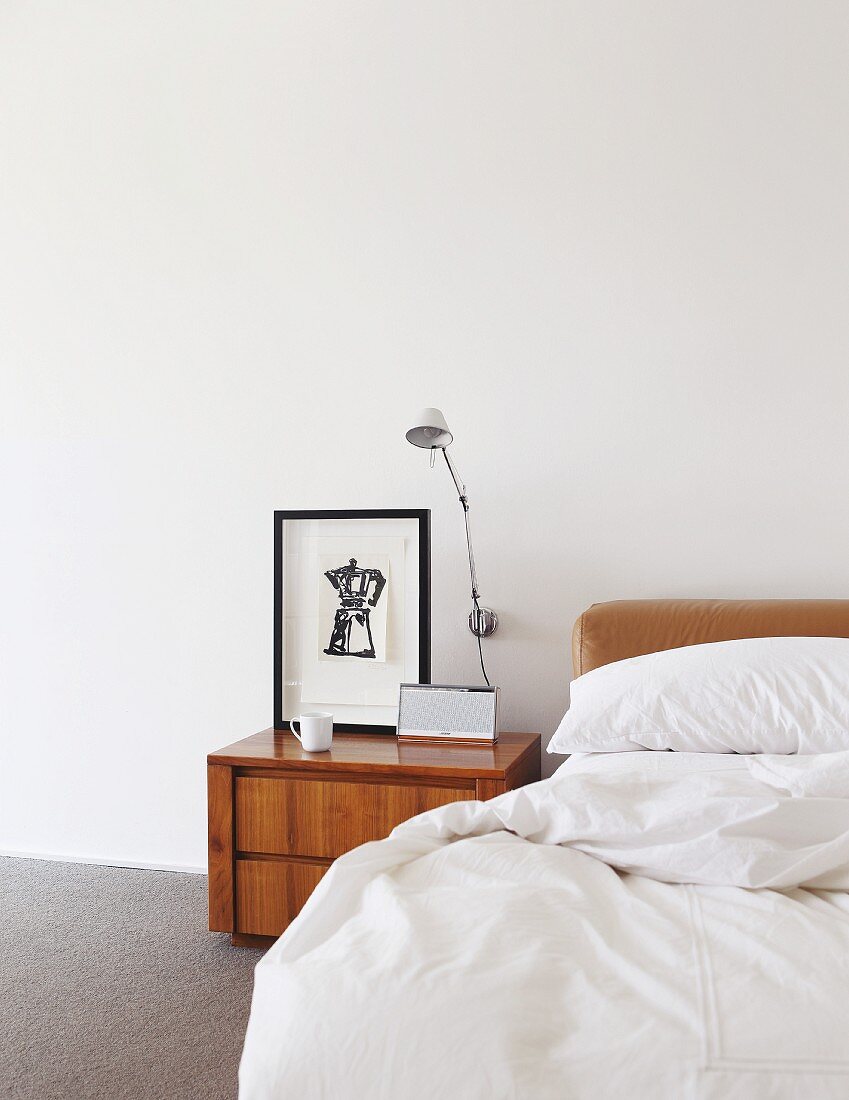Wooden bedside cabinet and wall-mounted lamp next to bed with white bed linen