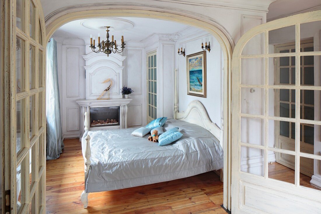 Offene Flügelsprossentür mit Blick in traditionelles Schlafzimmer mit glänzendem Plaid auf französischem Bett, im Hintergrund Kamin