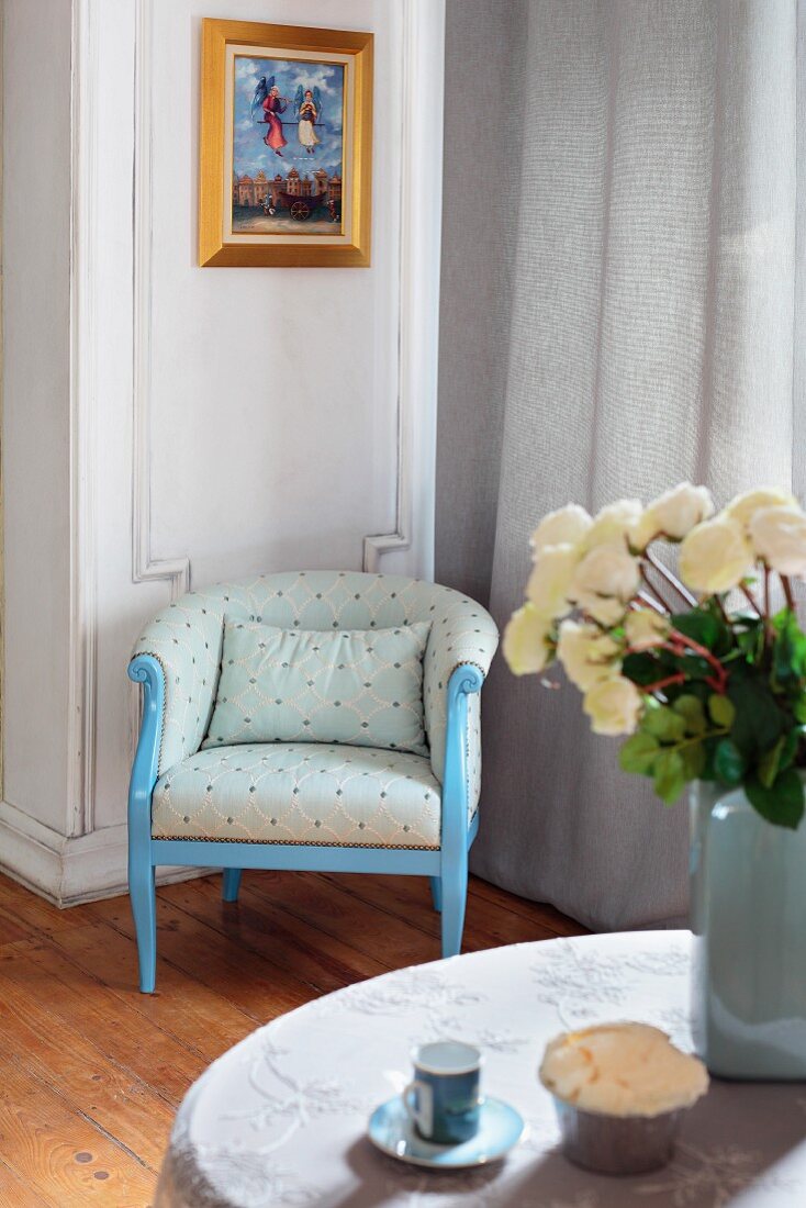 Blau lackierter Vintage Sessel mit hellem Stoffbezug in Wohnzimmerecke, neben bodenlangem Vorhang am Fenster, im Vordergrund weisser Rosenstrauss auf Tisch