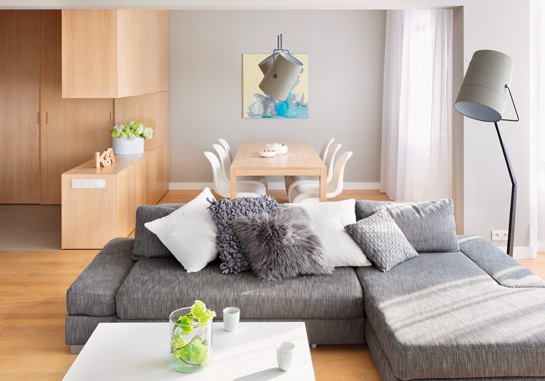 Graue Sofakombination mit Zierkissen, seitlich Scheinwerfer Stehleuchte in offenem Wohnraum mit Essplatz im Hintergrund