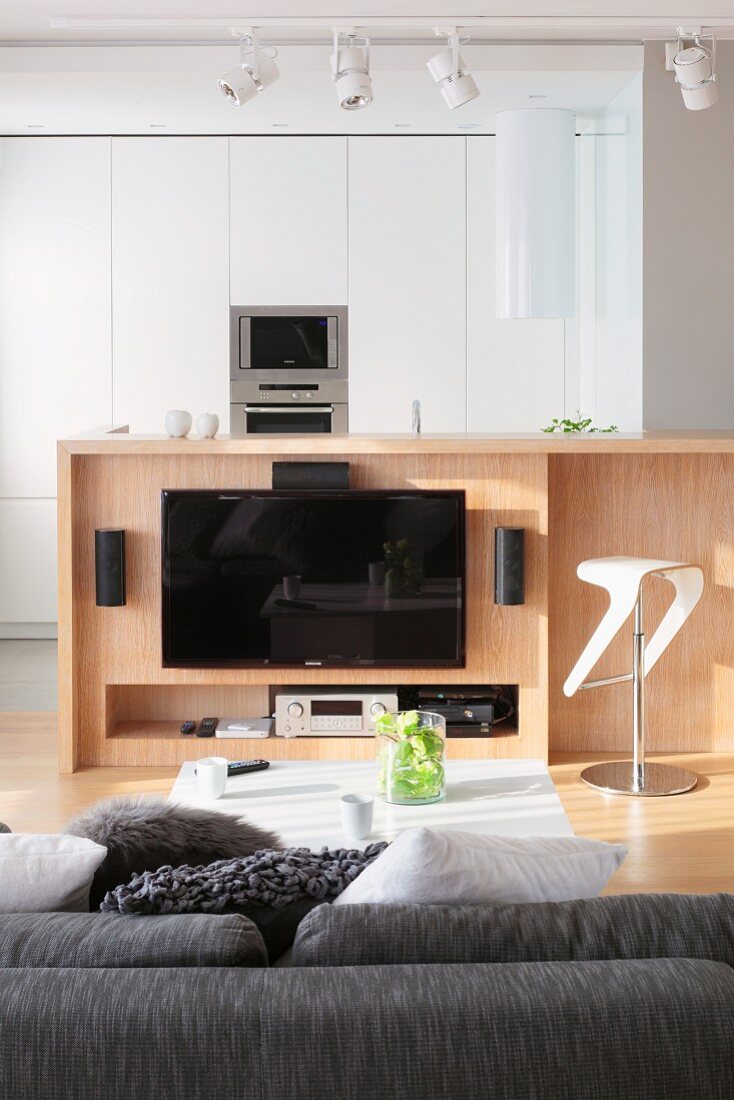 Blick vom Sofa auf Flachbildschirm an Holztheke montiert, im Hintergrund weisser Einbauschrank mit Küchengerät