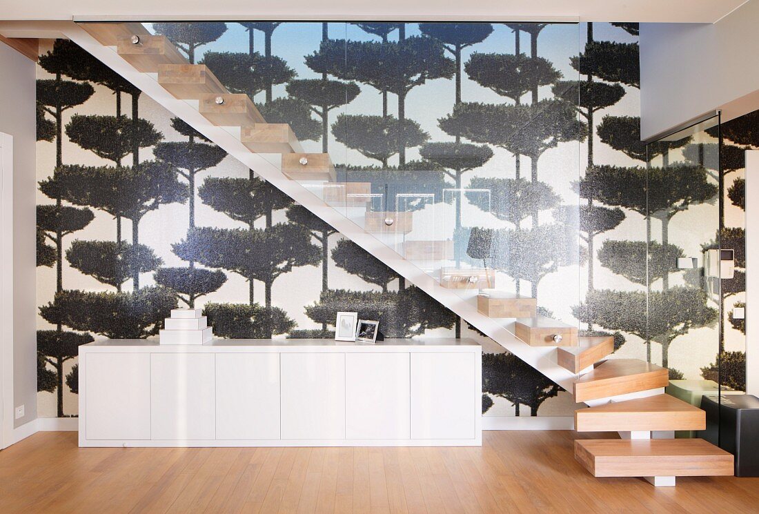 Weisses Sideboard unter Treppenaufgang mit Massivholzstufen auf Metallunterkonstruktion und Glasbrüstung, vor tapezierter Wand mit Baummotiven