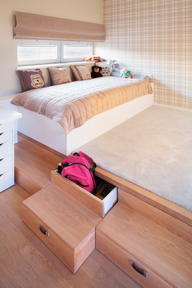 Platform with drawers in wooden steps, scatter cushions on child's bed and pale tartan wallpaper on wall