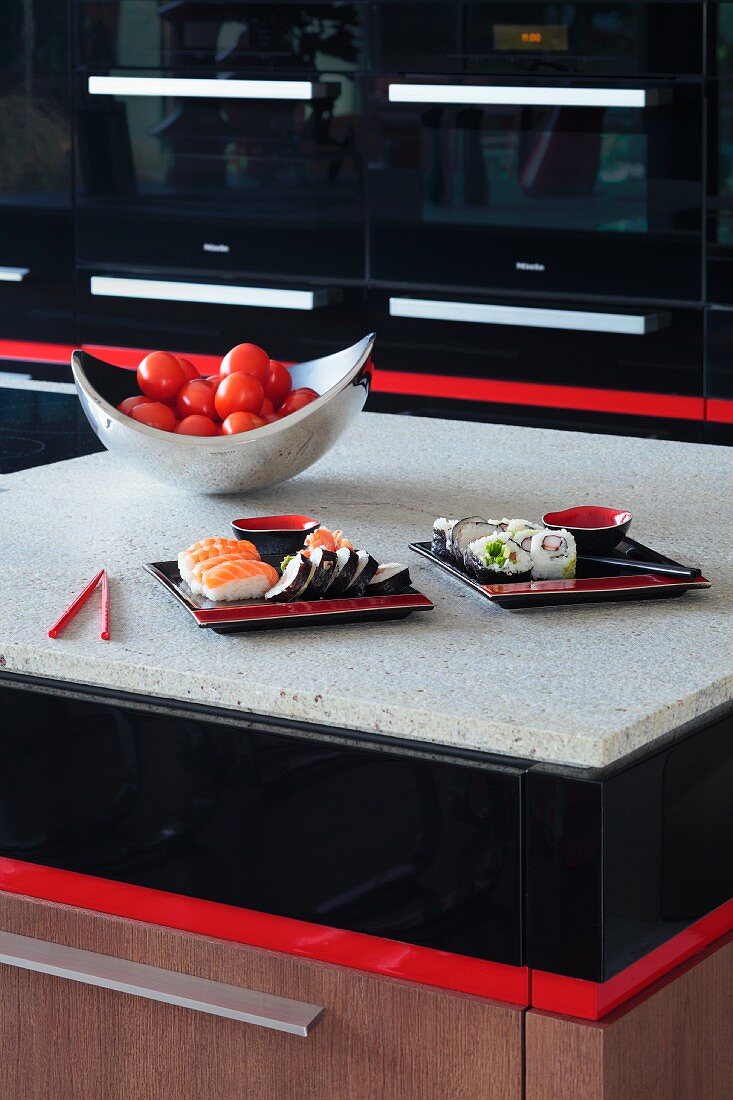 Detail of island counter with black and red accents, pale stone surface, bowl of tomatoes and two plates of sushi