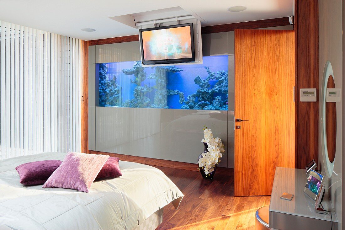 Bett mit Kissen in Violetttönen auf hellem Plaid, gegenüber Aquarium in Wand eingebaut, in modernem Schlafzimmer
