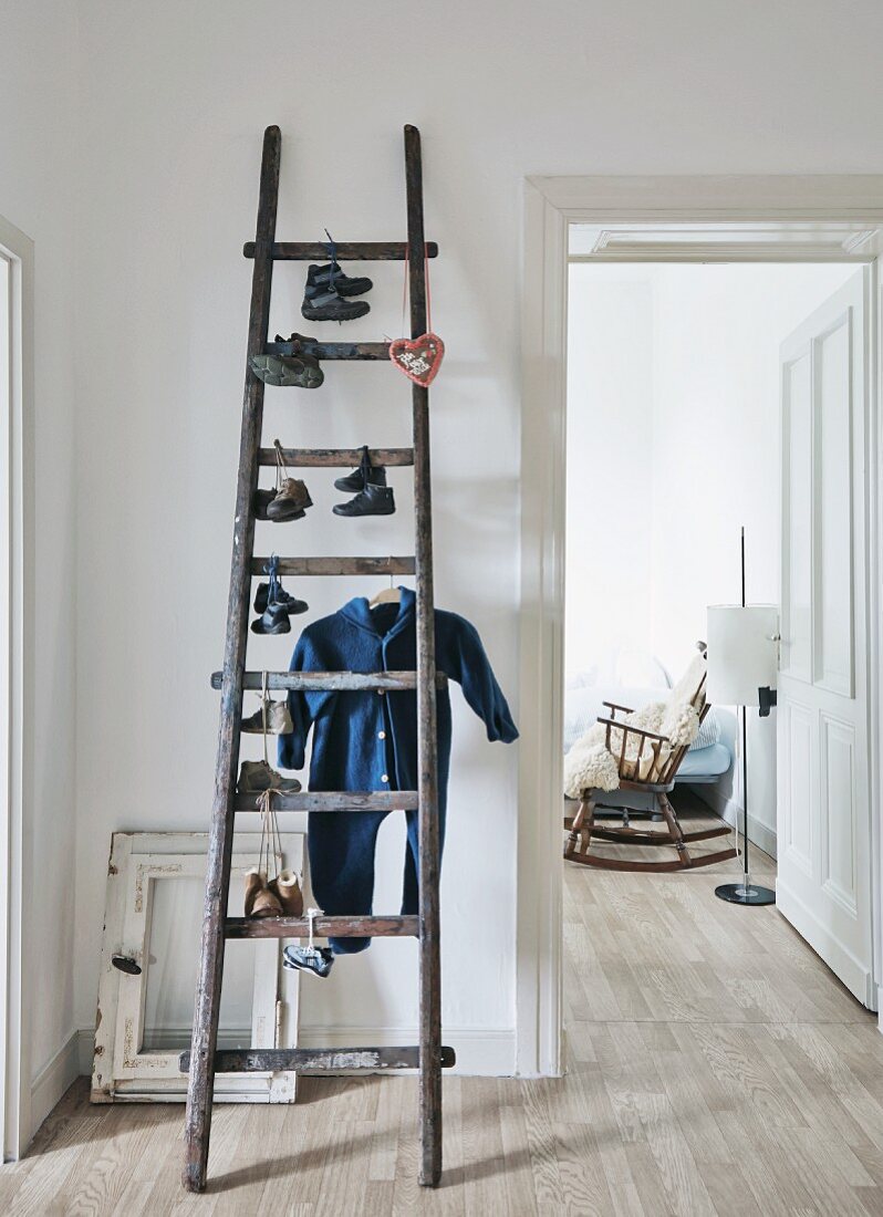Pairs of child's shoes and Babygro hanging from old attic ladder; old rocking chair seen through open door