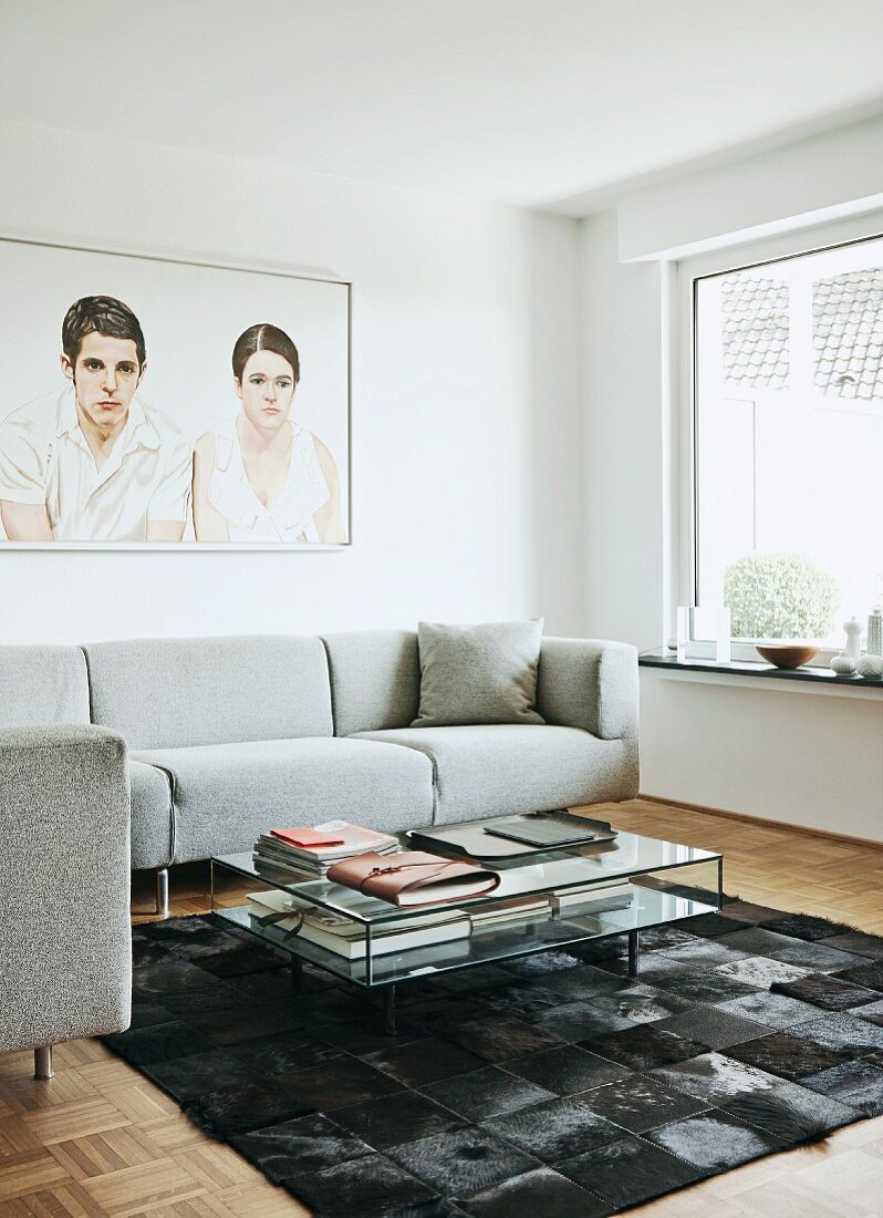 Coffeetable aus Glas auf Kuhfell Patchwork Teppich, elegante, moderne Couch in Hellgrau, vor Wandbild mit Männer- & Frauenmotv