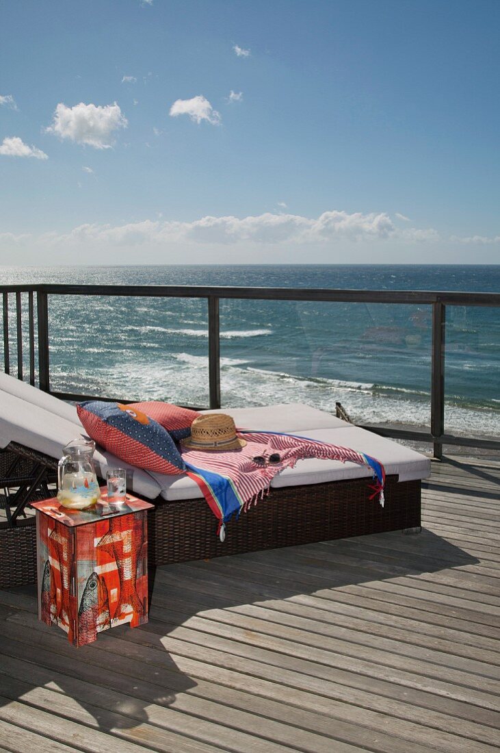 Gemütliche Sonnenliege mit Polsterauflage und bemalter Beistelltisch auf Holzdeck, Blick über Reling auf das Meer