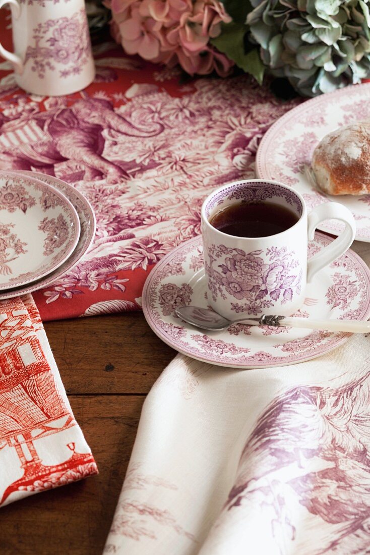 Geschirr und Tischwäsche mit rötlichen Toile-de-Jouy Mustern auf rustikalem Holztisch