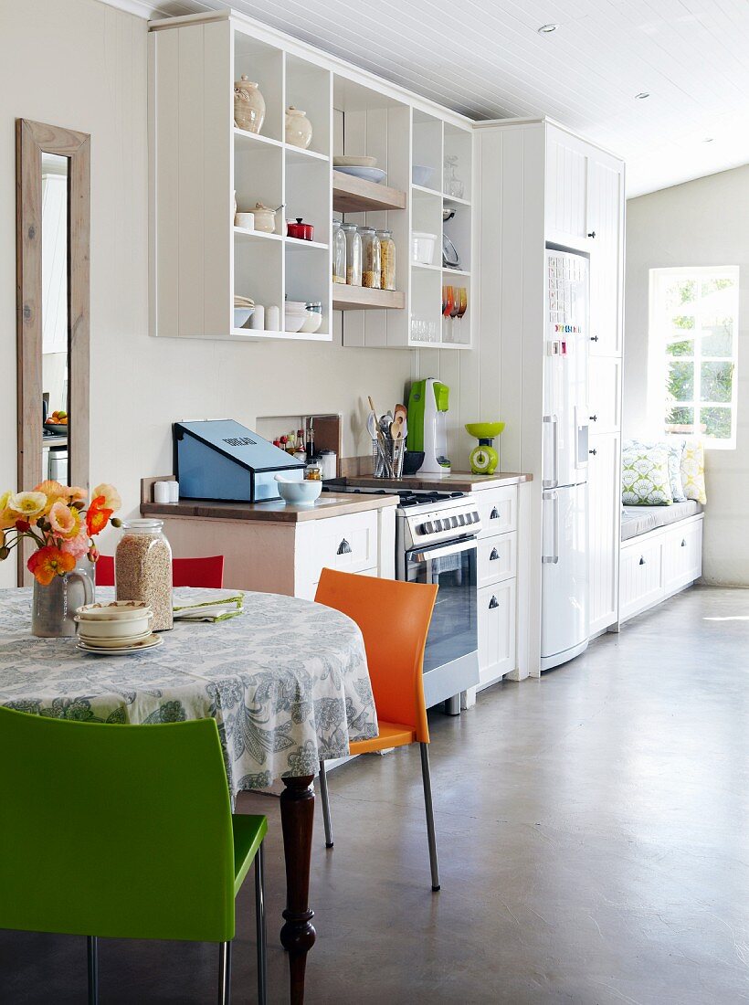 Bright, light-flooded kitchen with easy-care concrete floor, open-fronted shelves, round dining table with colourful chairs and bench with storage solution below window