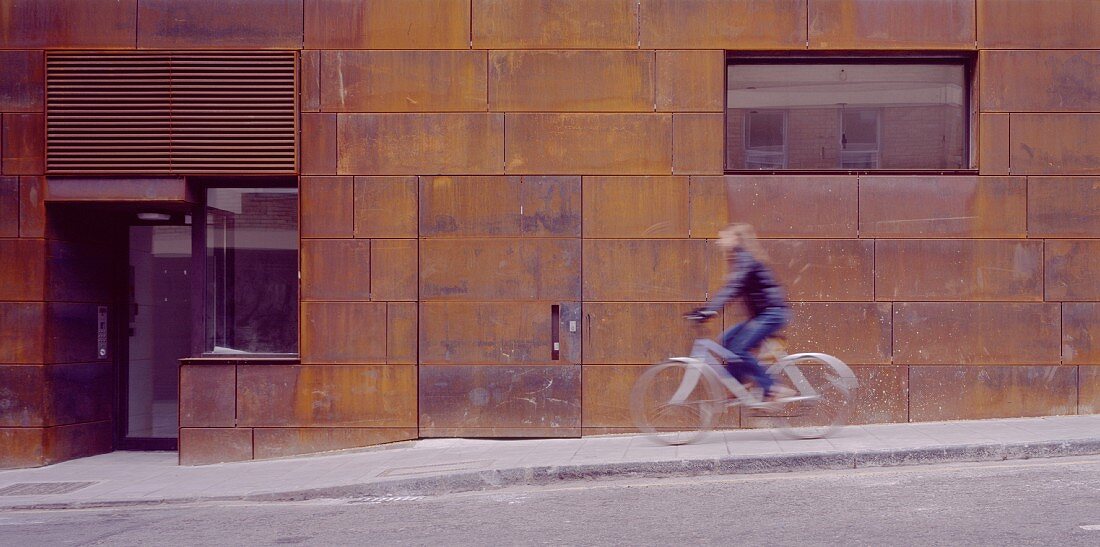 Lady riding a bike past a house facade with rusty Corten steel siding