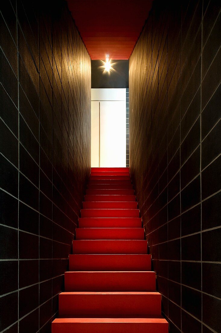 Schmaler Treppenaufgang in dramatischer Lichtinszenierung mit schwarzen Fliesenwänden und roter Treppe