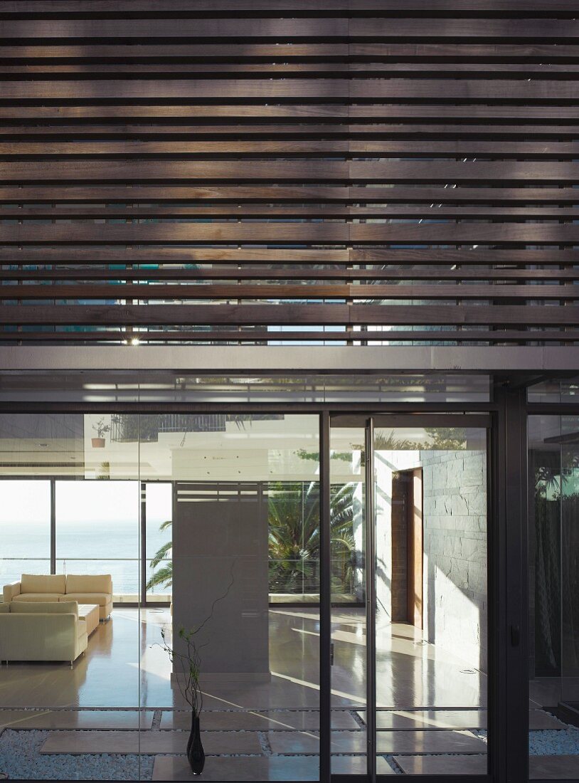 Moderne Neubauhausfassade mit Holzlamellen und verglastem Erdgeschoss mit Blick in minimalistischen Wohnraum