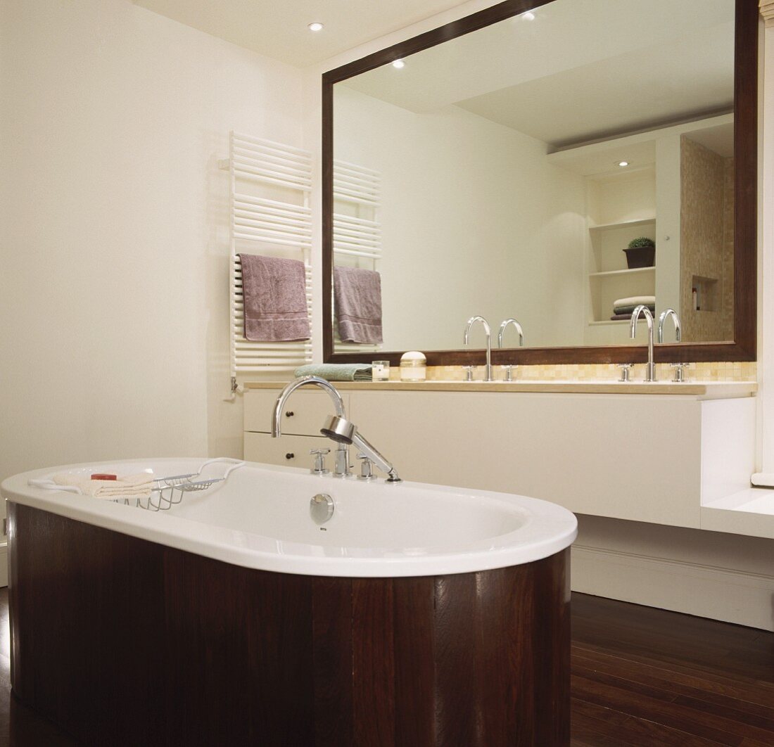 Freistehende Badewanne mit dunkler Holzverkleidung vor minimalistischem Waschtisch und Spiegel