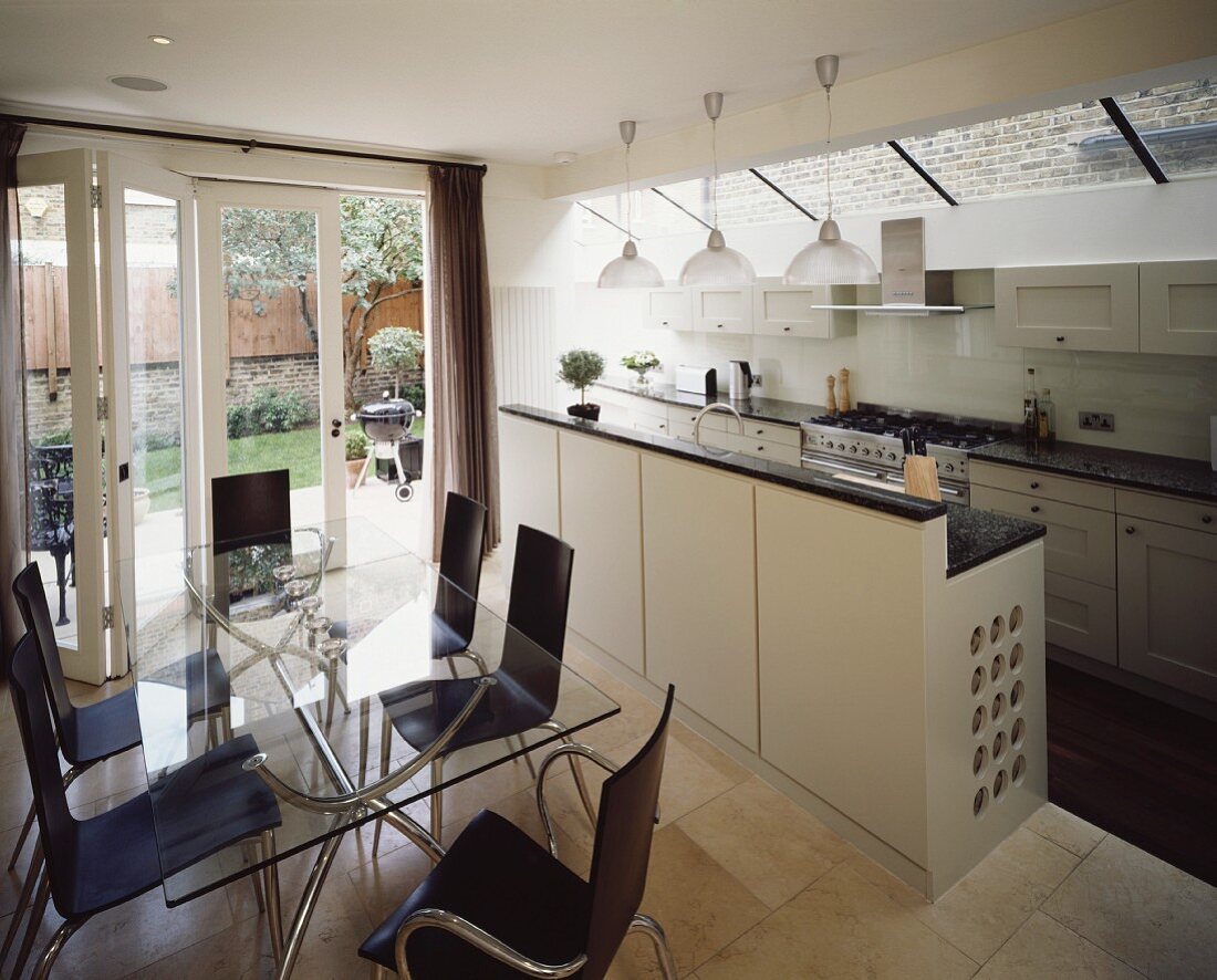 Moderne offene Küche mit weisser Theke und dunklen Holzschalenstühlen am Esstisch mit Glasplatte und Terrassentürfront