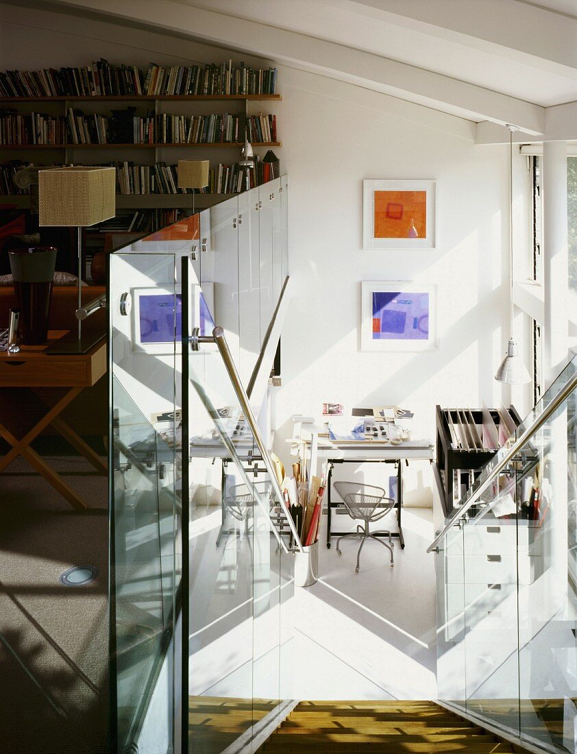 Descending stairs with a glass balustrade and view of a writing desk in an open room