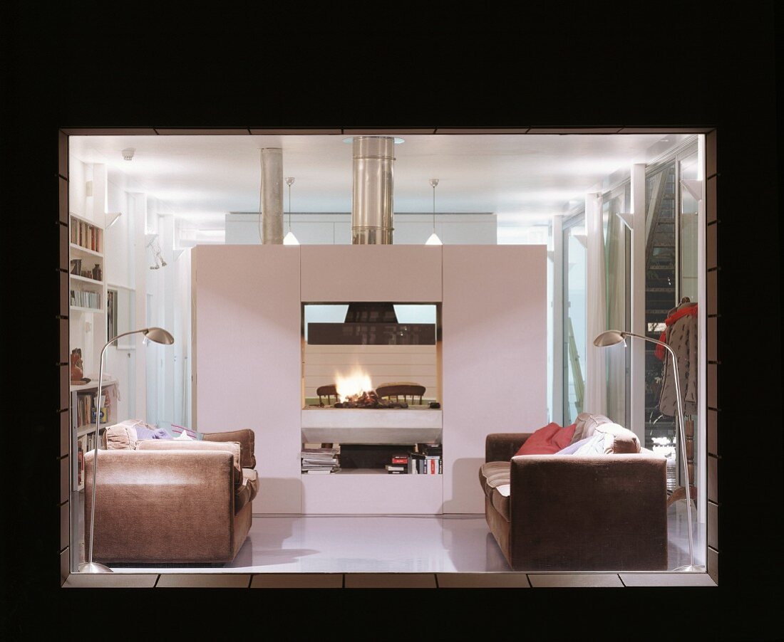 View through an illuminated window of a vintage leather sofa in front of a fireplace built into a free standing cube