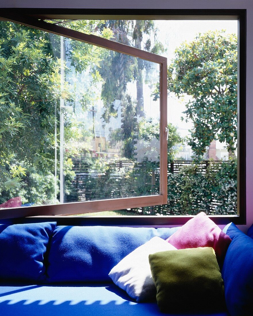 Sofa mit blauem Bezug und farbigen Kissen vor offenstehendem Fenster und Gartenblick