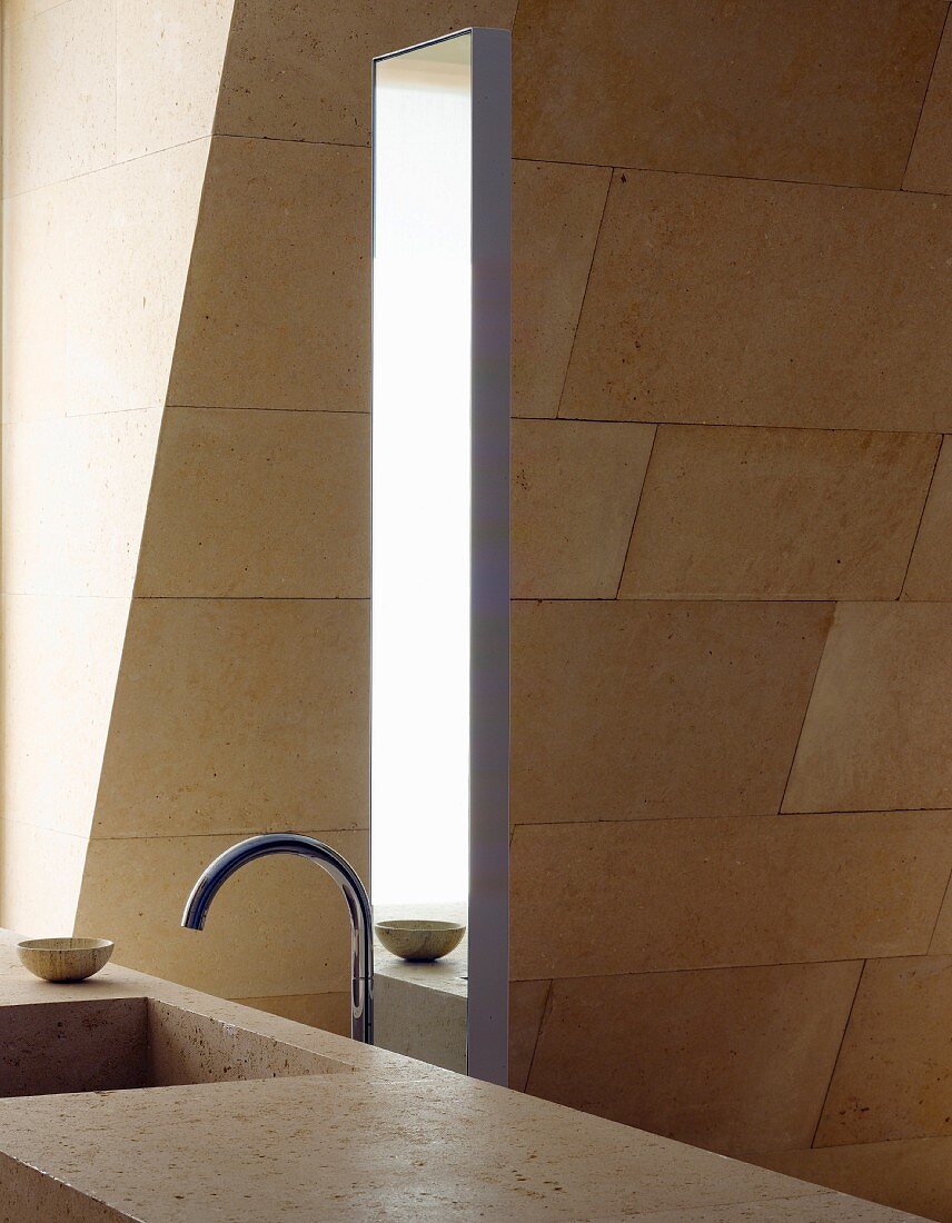 Ausschnitt eines Steintisches mit Becken und Designerarmatur vor Spiegel und Steinfliesen an Wand